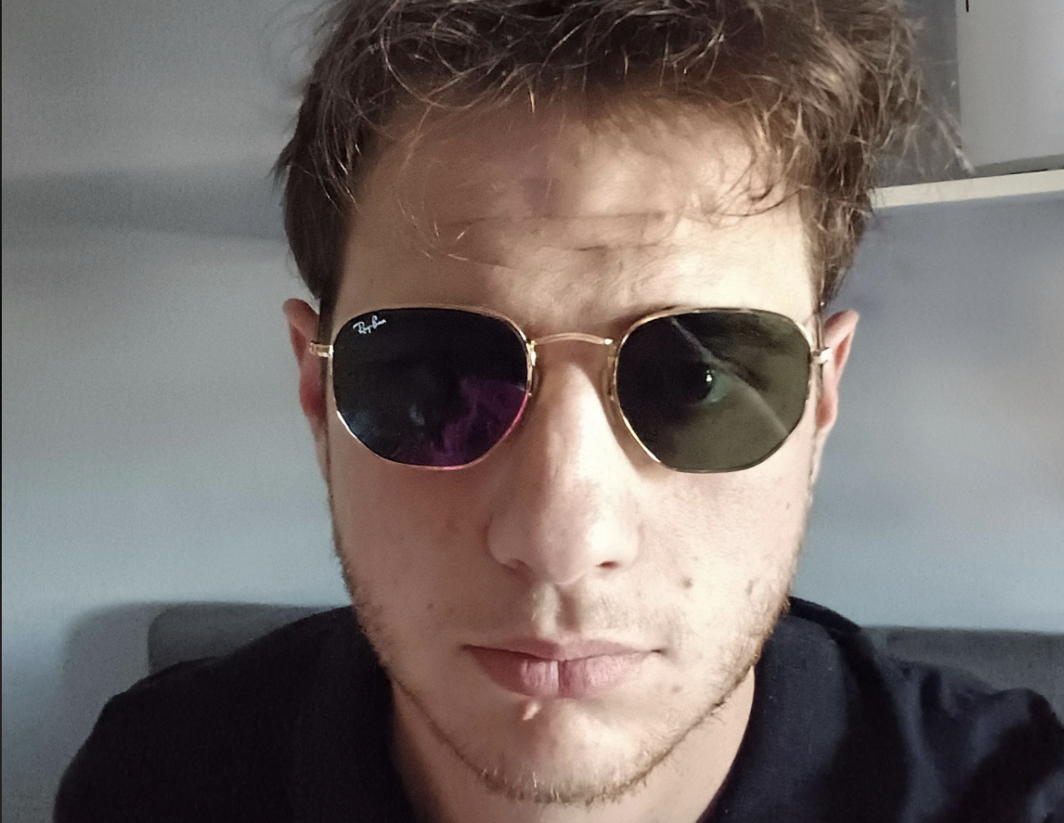 A young man with short brown hair and light facial hair wears dark aviator sunglasses and a black shirt, looking directly at the camera in a softly lit indoor setting.