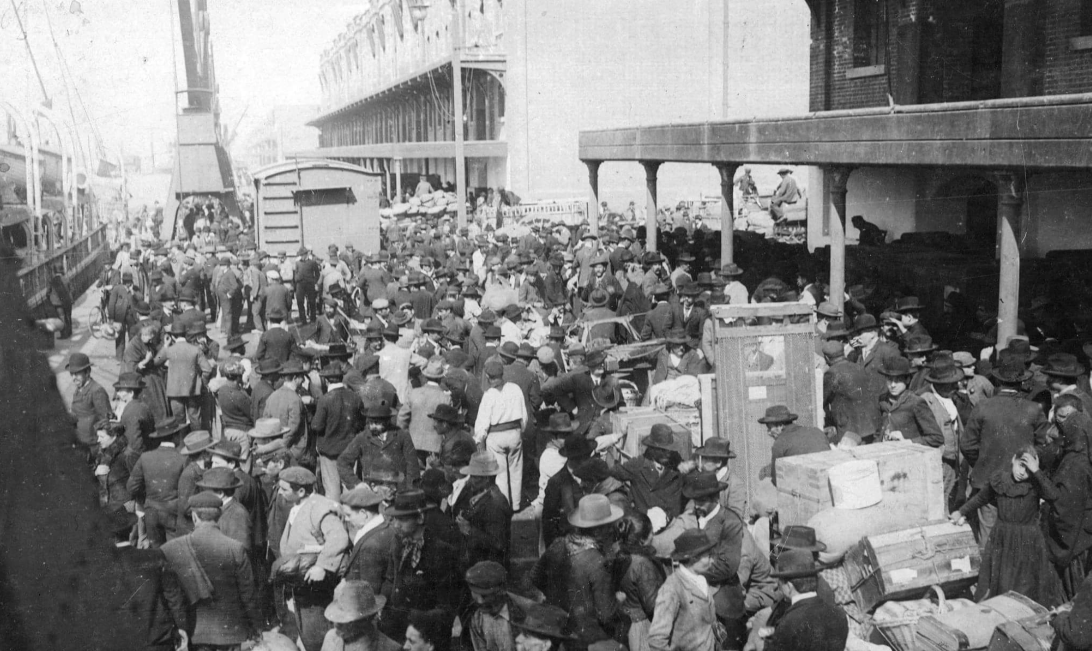 A large crowd of people, many wearing hats and coats, gather with luggage and belongings on a busy dock beside a building, likely waiting to board or having arrived by ship. The scene appears historical and bustling.