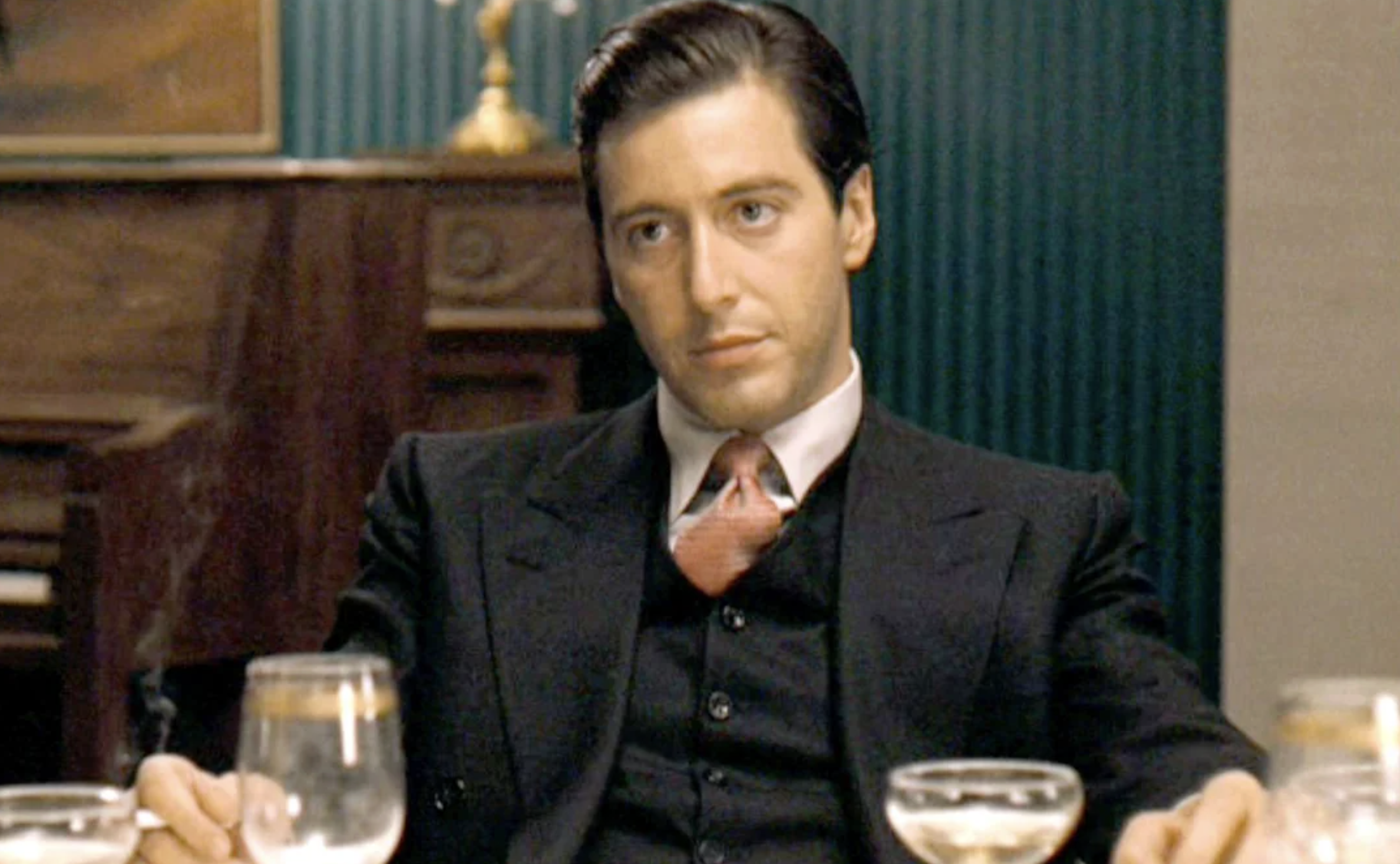 A man in a black suit and pink tie sits at a table with glassware, looking serious and composed. The background features a piano and green vertical-striped walls.