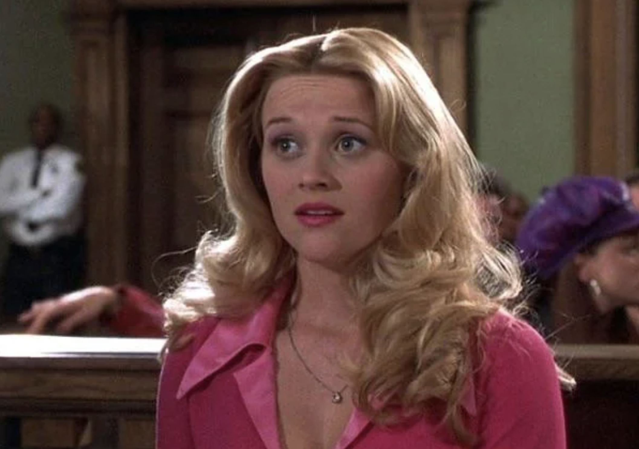 A blonde woman wearing a pink outfit sits in a courtroom, looking surprised or concerned. There are people and a uniformed guard in the background.