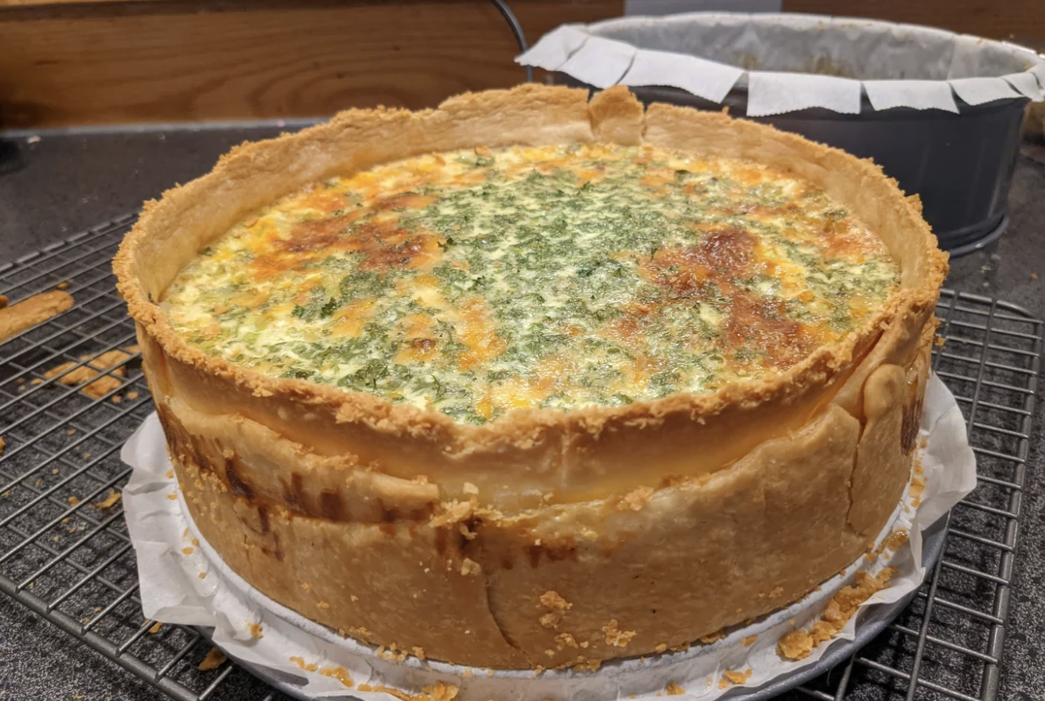 A freshly baked quiche with a golden, thick crust and a creamy filling topped with herbs, resting on a cooling rack with parchment paper underneath. Another baking pan is visible in the background.