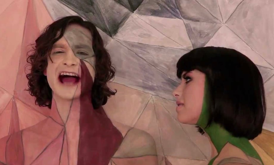 Two people with geometric patterns painted on their faces and bodies stand against a matching abstract background. One person is singing with mouth open while the other, facing sideways, looks at them.