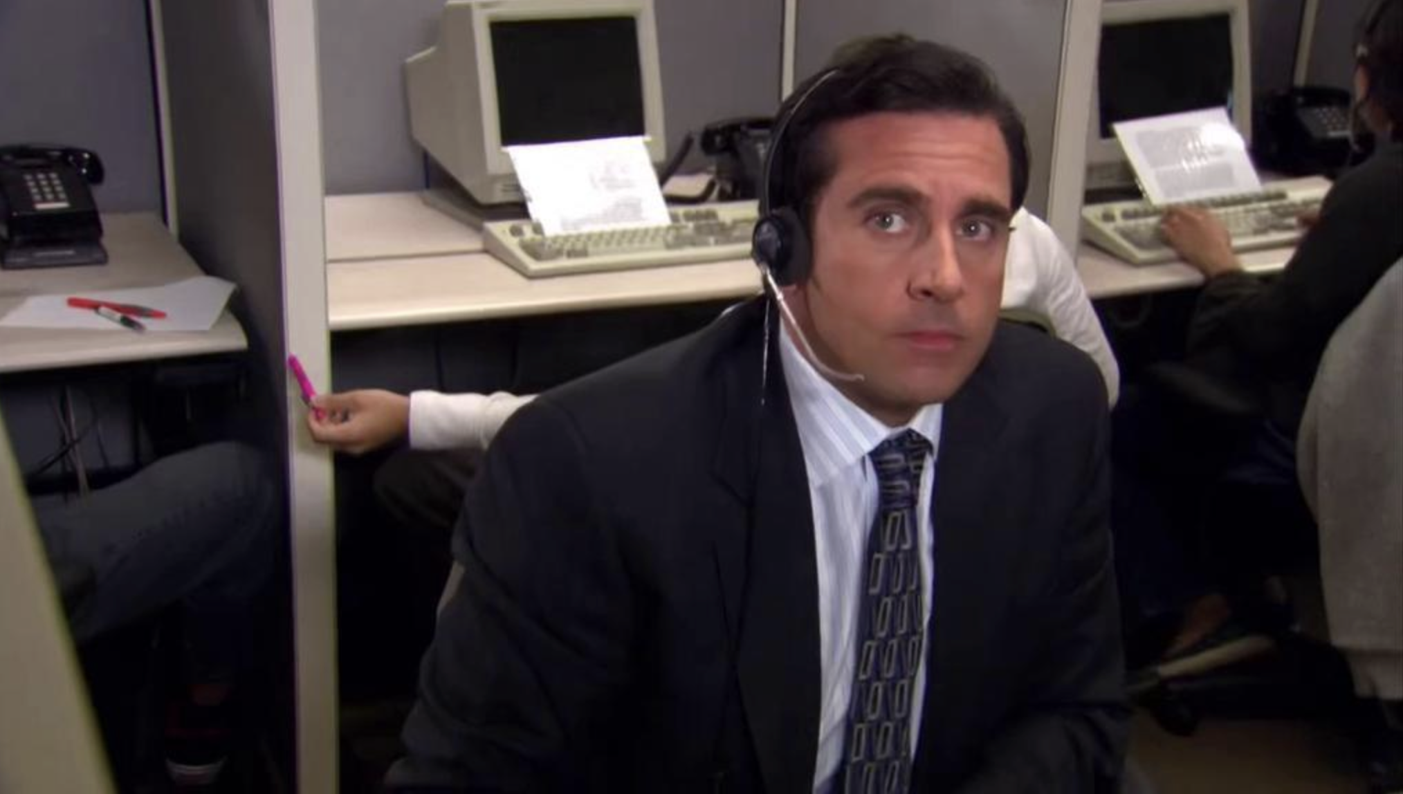 A man in a suit and tie wears a headset and sits in an office cubicle, looking at the camera. Behind him, coworkers work at computers in separate cubicles. One hand reaches out, passing a pink highlighter.