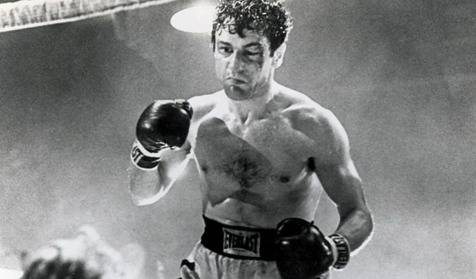 A black-and-white photo of a male boxer in a boxing ring, wearing boxing gloves and shorts. He looks focused and sweaty, with curly hair and his hands raised, ready to fight. An audience or opponent is partly visible.