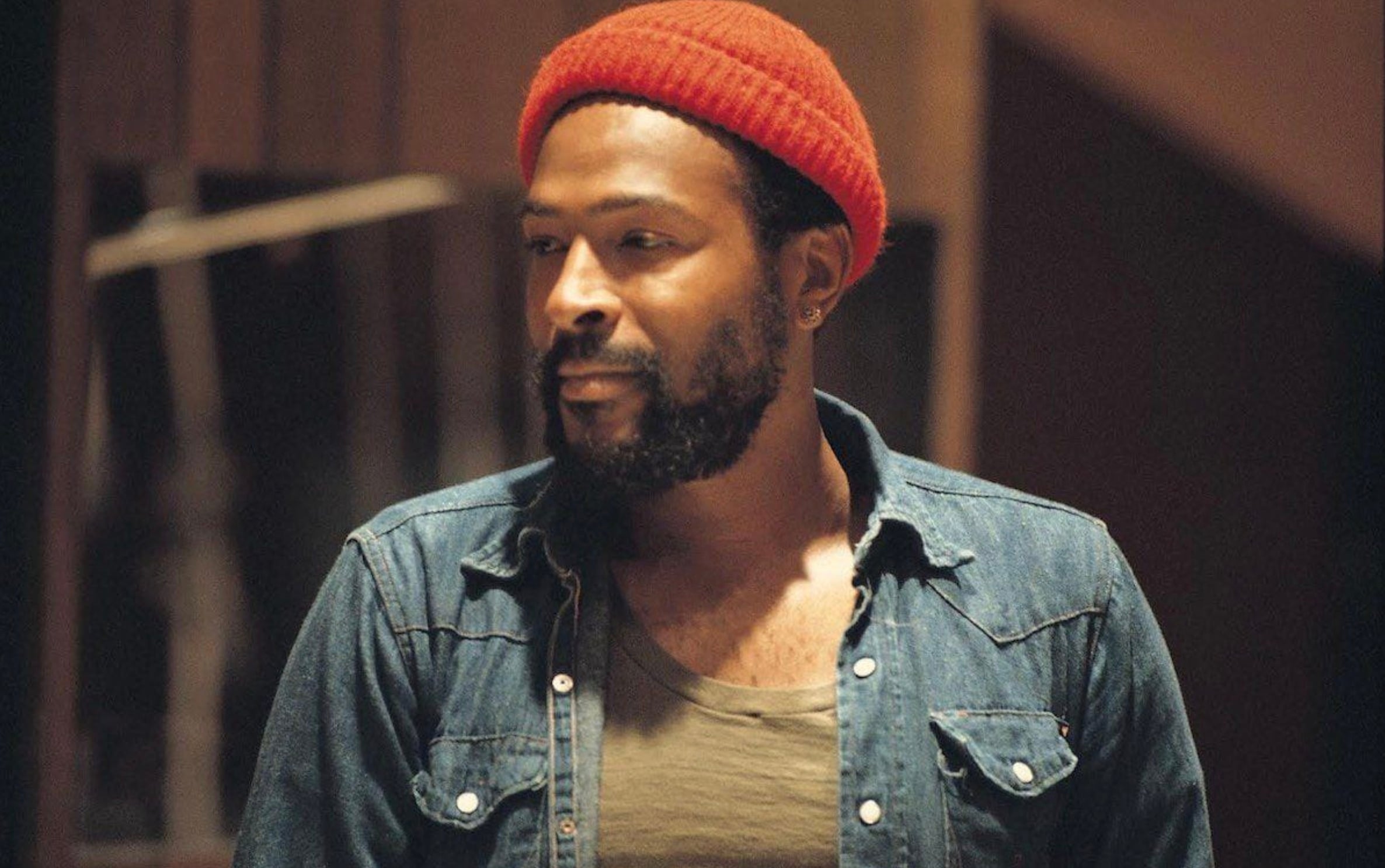 A man with a beard wears a red knit hat, olive green shirt, and an unbuttoned blue denim shirt, looking to the side in a softly lit indoor setting.