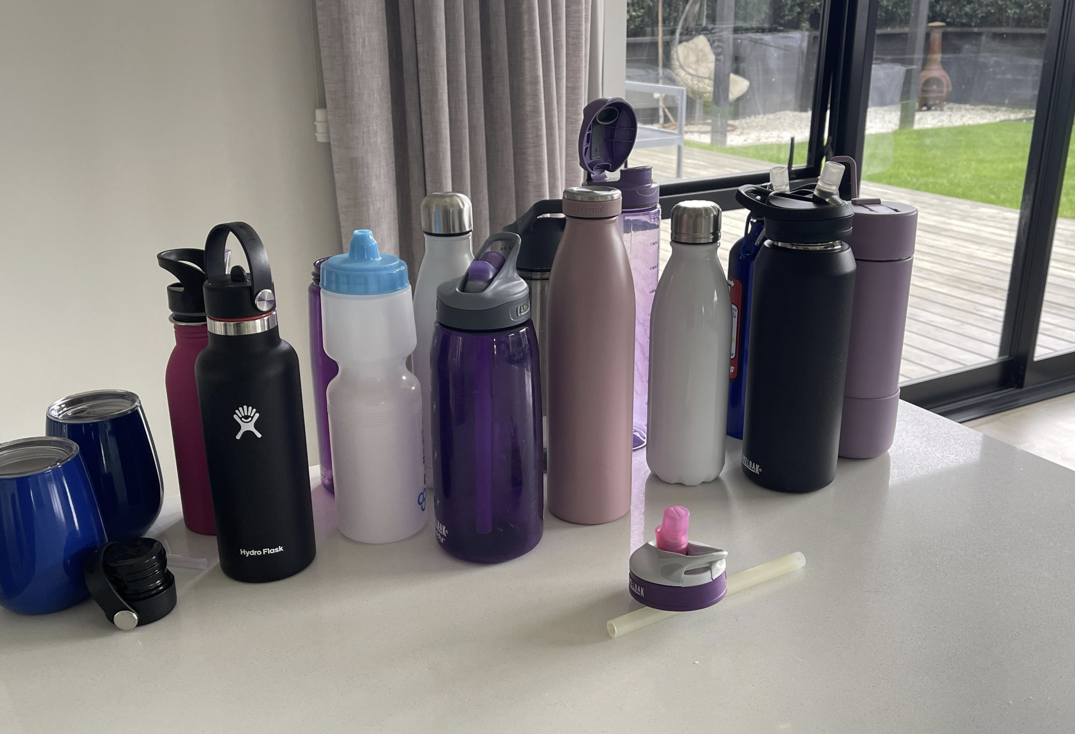 A collection of reusable drink bottles and tumblers in various colors and sizes are arranged on a white kitchen counter near a window with a view of a patio and garden outside.