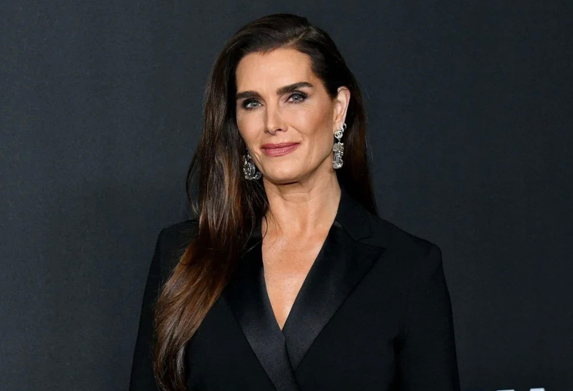 A woman with long brown hair wearing a black suit jacket and large dangling earrings stands in front of a dark background, smiling softly at the camera.