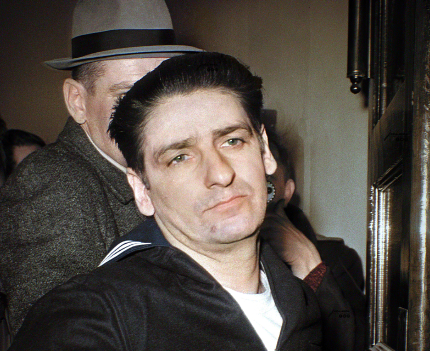 A man with dark hair and a somber expression looks slightly past the camera. He wears a dark jacket and stands in front of another man in a gray suit and hat, with a crowd and doorway in the background.