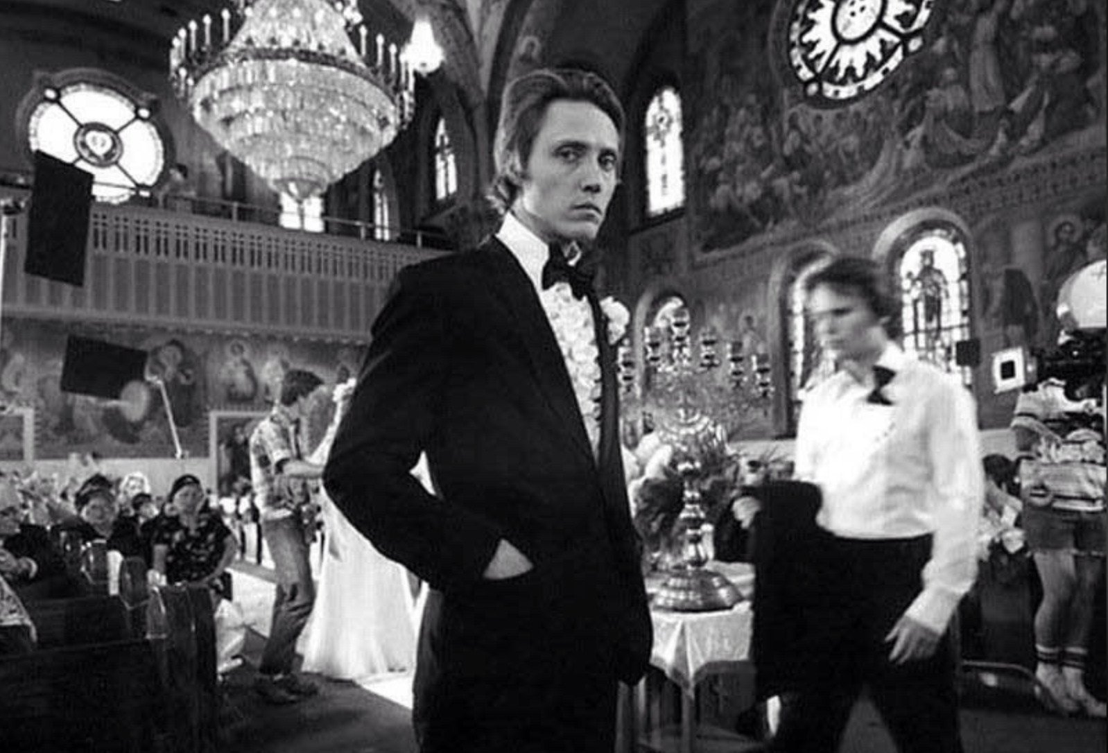 A man in a tuxedo stands with hands in his pockets inside an ornate church, with people and film equipment visible in the background, suggesting a wedding scene being filmed.