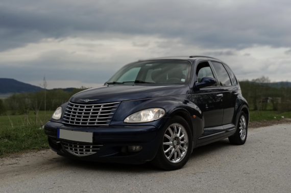 A dark blue Chrysler PT Cruiser is parked on a rural road, with grassy fields and cloudy skies in the background.