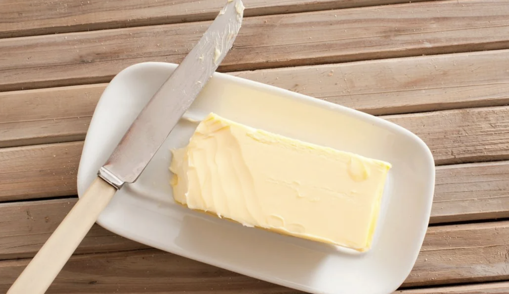 A stick of butter with a few slices missing sits on a white dish. A butter knife with some butter residue rests next to it. The dish is placed on a wooden surface.