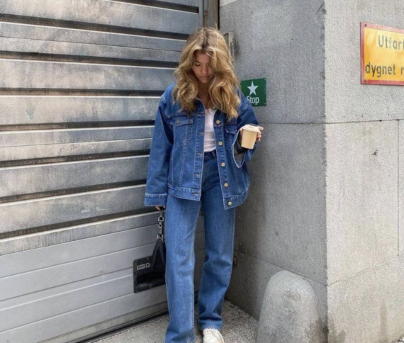 A woman with wavy hair stands by a gray garage door and concrete wall, wearing a blue denim jacket and jeans, holding a black handbag and a paper coffee cup, and looking down.