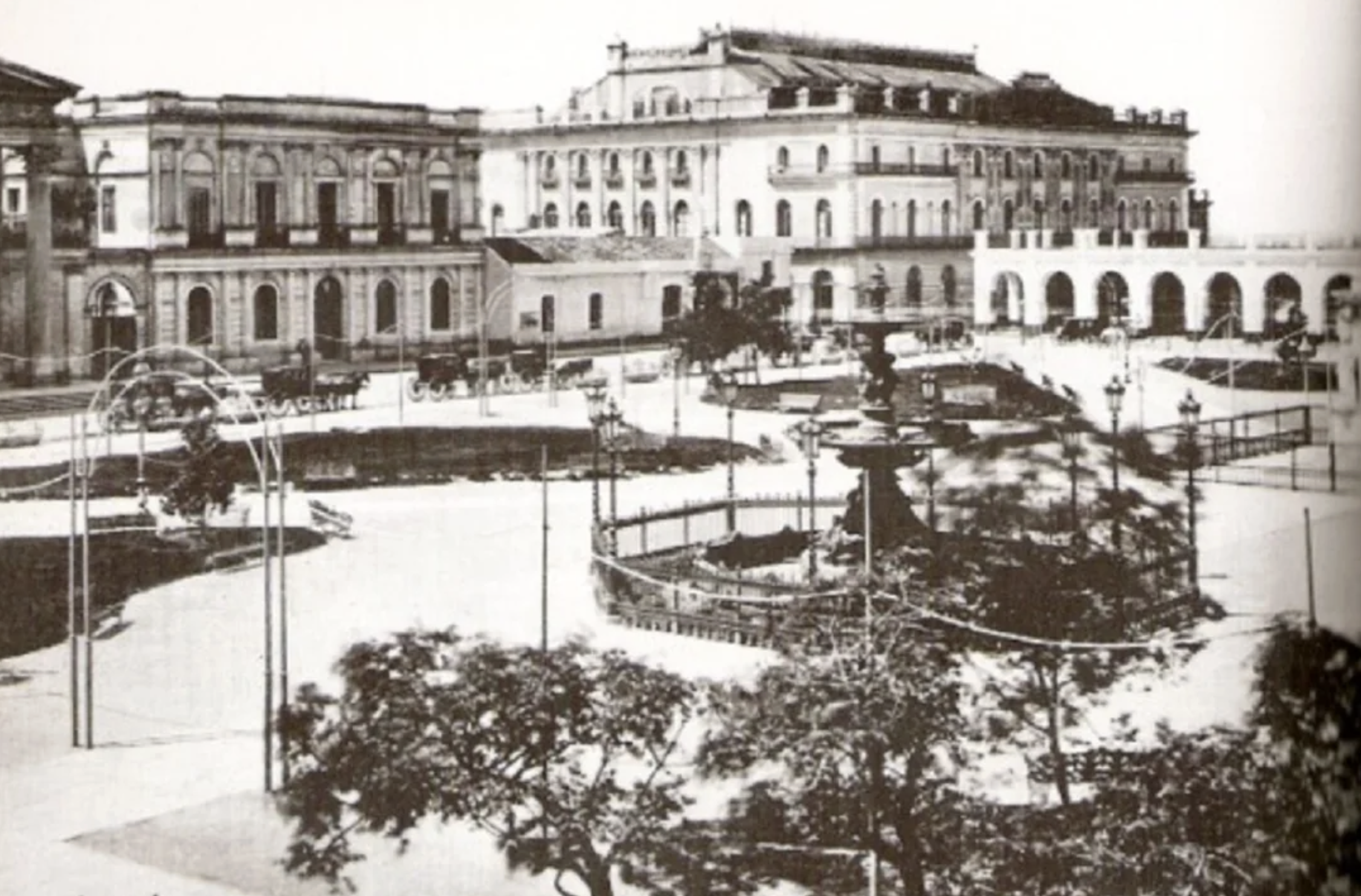 Historic black and white photo of a city square with trees, a central fountain, a gazebo, and ornate buildings in the background, showing classic architecture and a peaceful atmosphere.