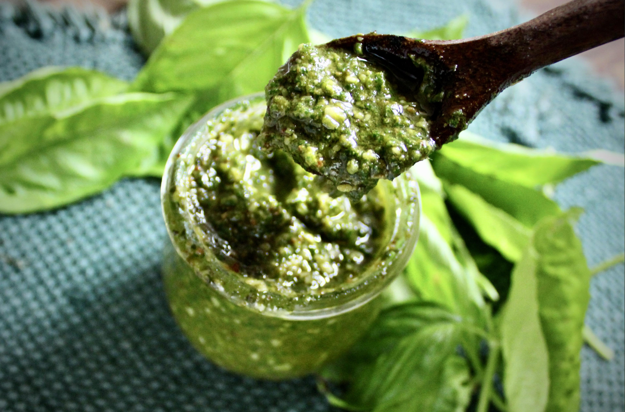 A wooden spoon holds a scoop of green pesto over a glass jar, with fresh basil leaves scattered on a textured blue surface in the background.