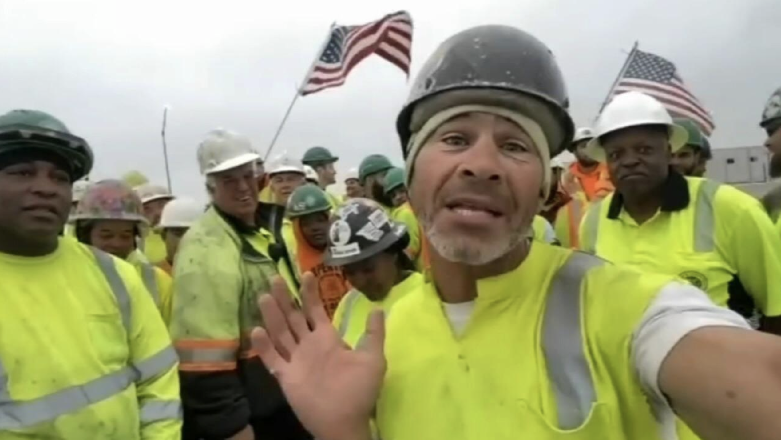 A group of construction workers in bright yellow safety gear and helmets pose together outdoors. Two American flags are visible in the background. One worker is at the front, gesturing towards the camera.