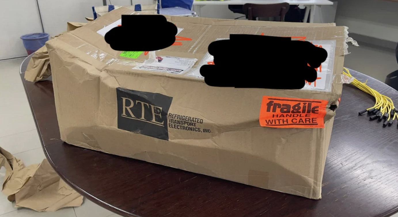 A large cardboard box labeled "fragile" and "RTE" sits on a dark table. The box is crumpled and damaged, with shipping labels partially obscured. Cables and packing materials are nearby.