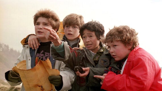 Four boys stand outdoors, closely huddled together. One points ahead urgently while another holds a worn treasure map. They all look intently in the same direction, appearing excited and determined.
