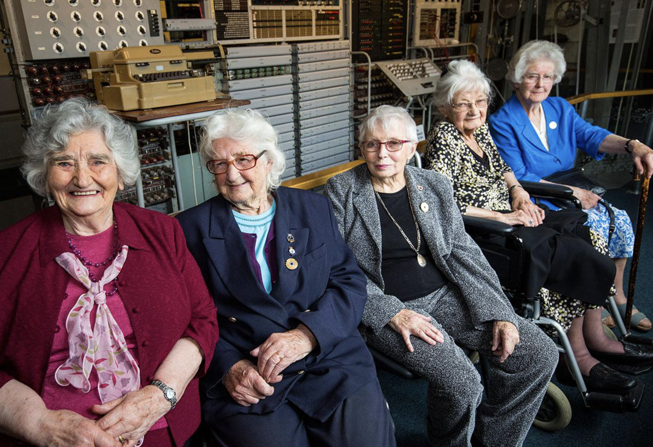 Five elderly women, smiling and seated in front of vintage computers and machinery, appear to be in a museum or historical setting, wearing formal attire and badges.