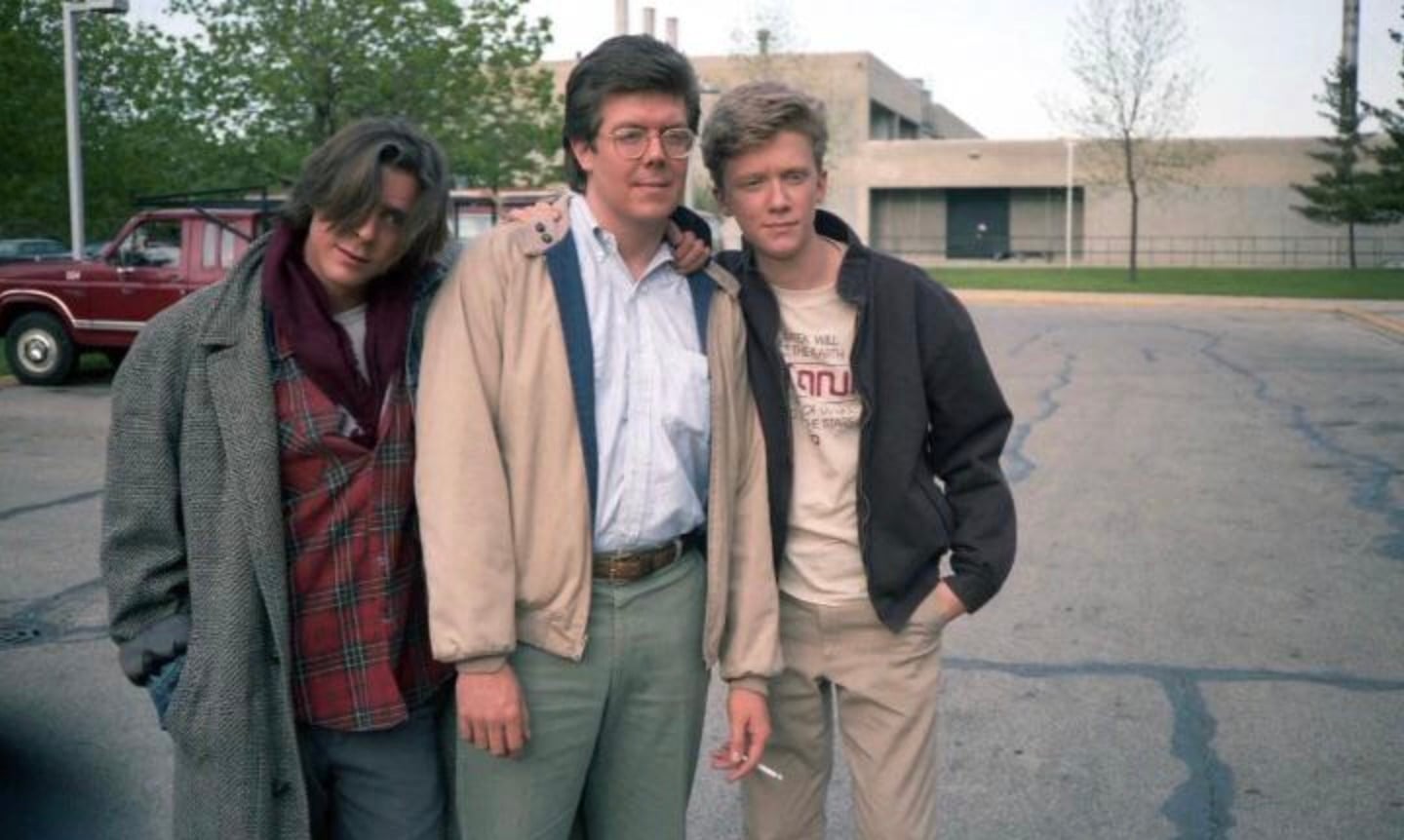 Three people stand together in a parking lot, posing for a photo. Two young men lean casually on an older man in the middle. There are trees, a red truck, and a building in the background.