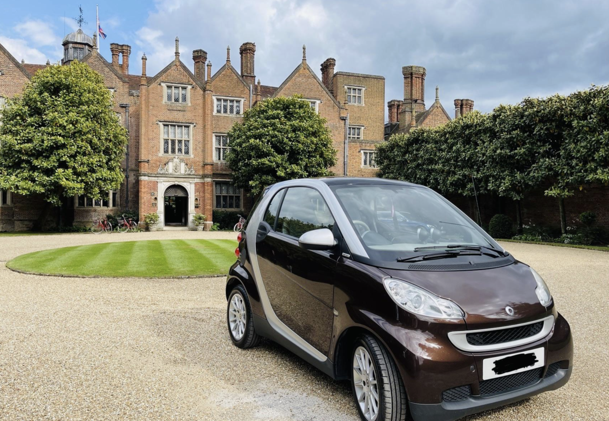 A brown Smart car is parked on a gravel driveway in front of a large, historic brick mansion with chimneys and lush green trees.