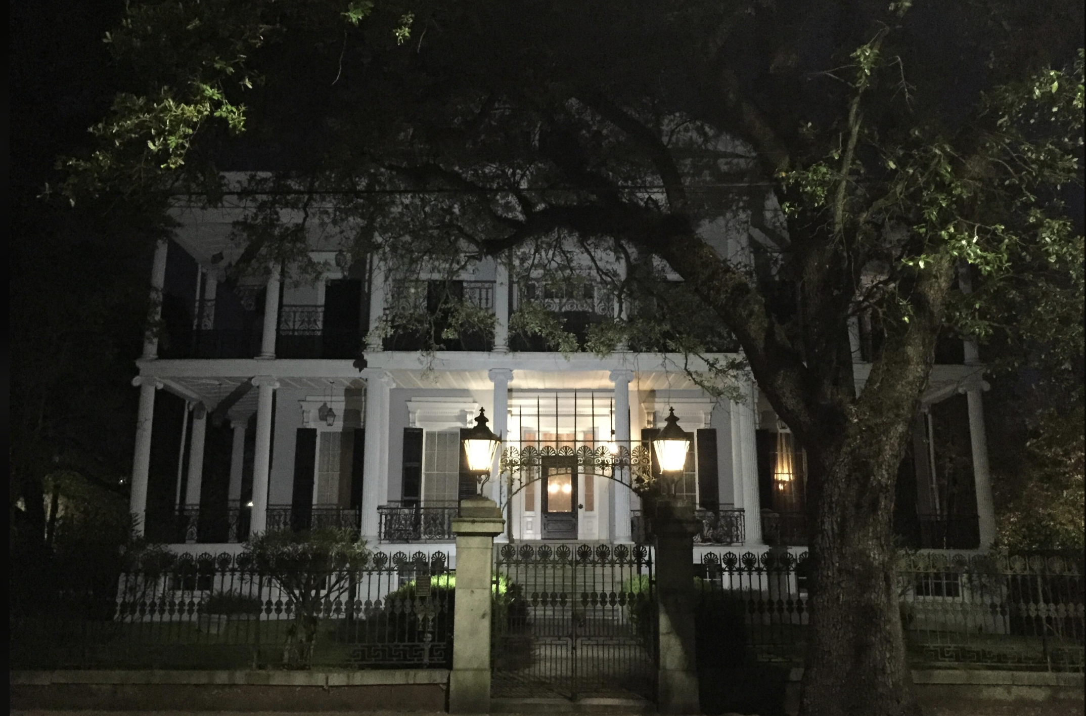 A large, two-story white house with tall columns and wrought iron balconies is lit up at night. Two lanterns stand on stone pillars by a gated entrance, and a large tree partially obscures the view.