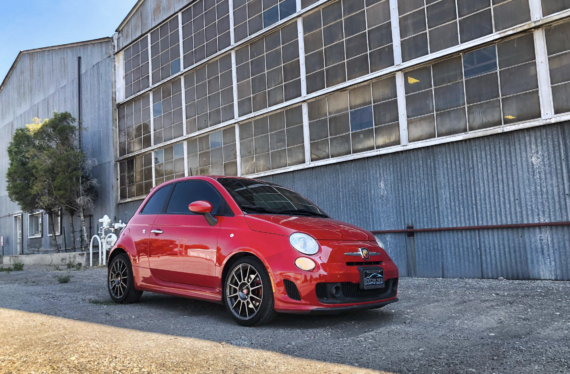 A bright red compact car is parked on a gravel surface in front of a large industrial building with metal siding and many tall windows. The sky is clear and blue.