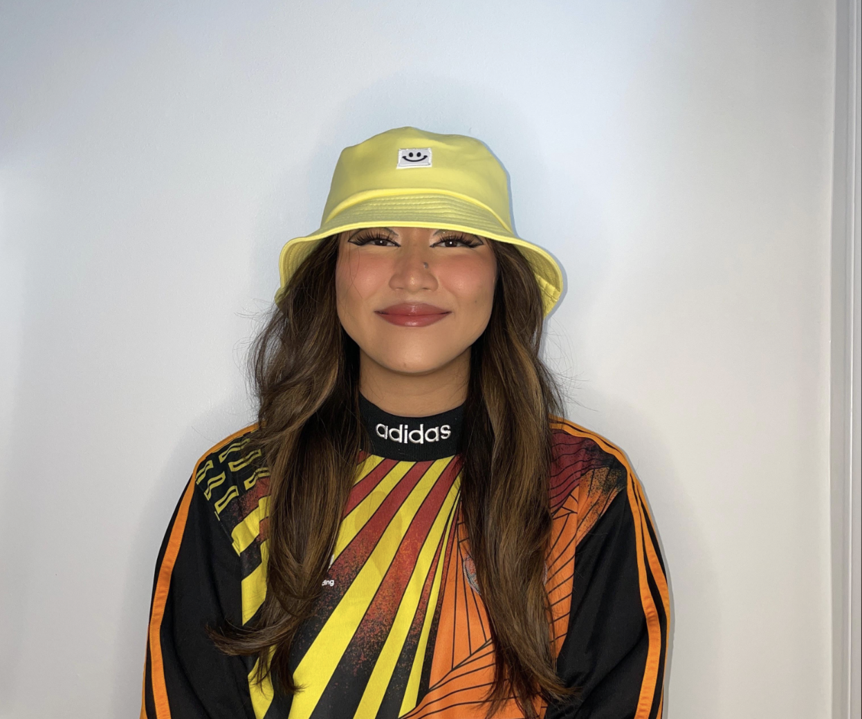 A smiling woman with long brown hair wears a yellow bucket hat, a black Adidas shirt with a bold yellow and red graphic, and stands against a plain white background.