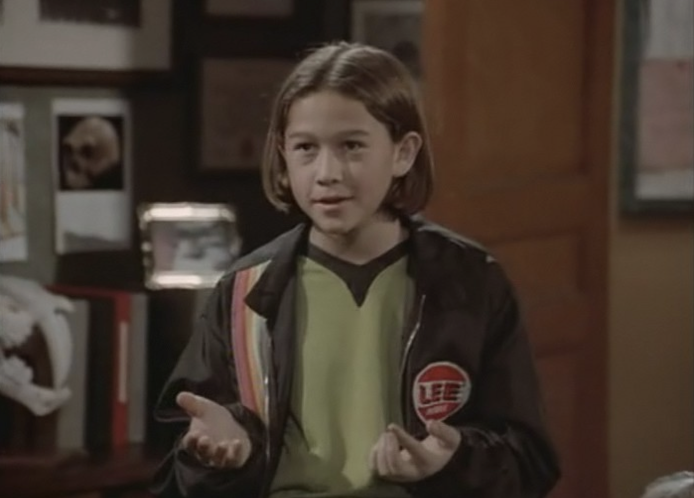 A young person with straight, shoulder-length hair stands indoors wearing a black jacket with a red "LEE" patch and a green shirt, gesturing with both hands while talking.