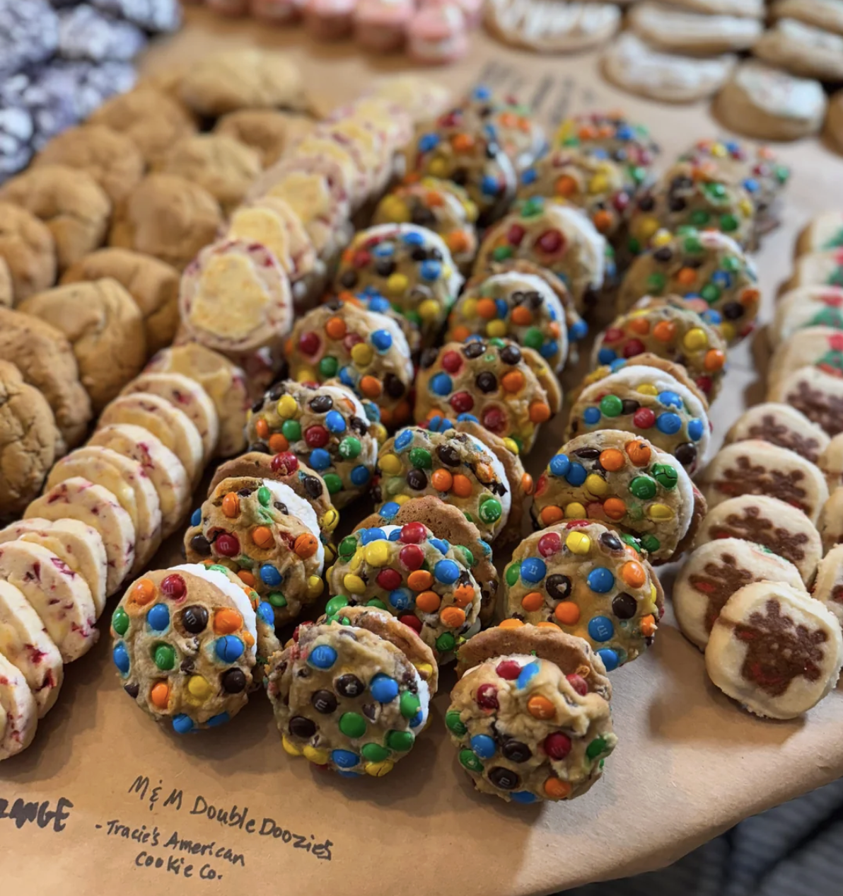 Rows of assorted cookies are displayed on brown parchment paper. In the center are cookie sandwiches with colorful M&Ms, surrounded by various types of cookies, including ones with white icing and red specks.