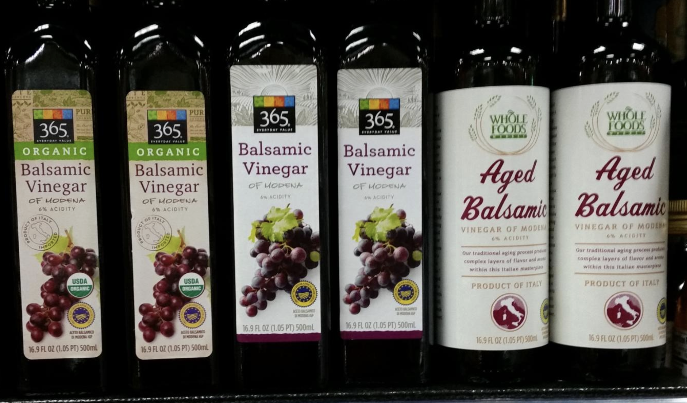 Five bottles of balsamic vinegar from Whole Foods' 365 brand and Whole Foods Market; labels feature grapes and text, with organic and aged varieties displayed on a store shelf.