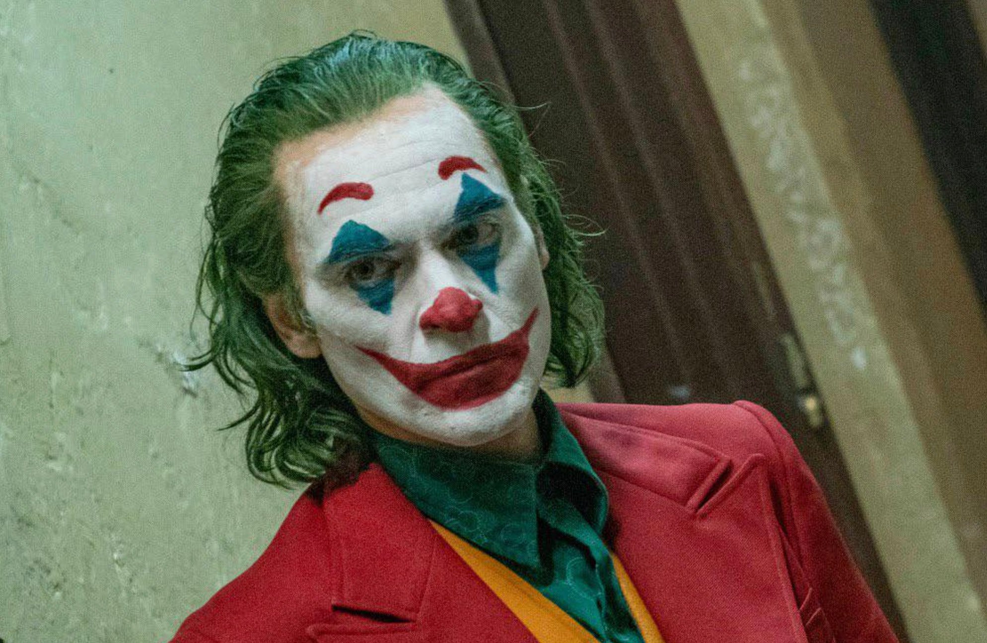 A person dressed as a clown with white face paint, red lips, blue eye makeup, green hair, and wearing a red suit jacket over a green shirt, stands indoors against a beige wall and wooden door.