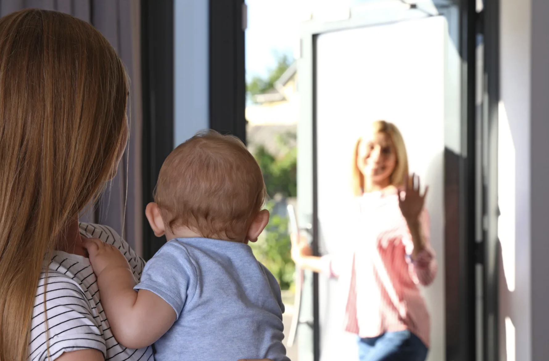 A woman holding a baby stands inside a house, looking toward another woman who is waving and entering through the open front door on a sunny day.