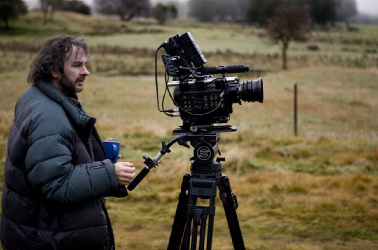 A man in a puffy jacket stands outdoors in a grassy field, holding a blue mug and operating a large professional video camera on a tripod, with trees and fog in the background.