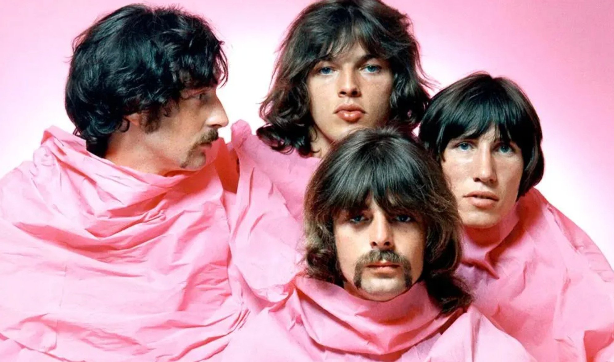 Four men with shaggy hair and mustaches, wrapped in pink cloth, pose closely together against a pink background. Their expressions are serious and contemplative.