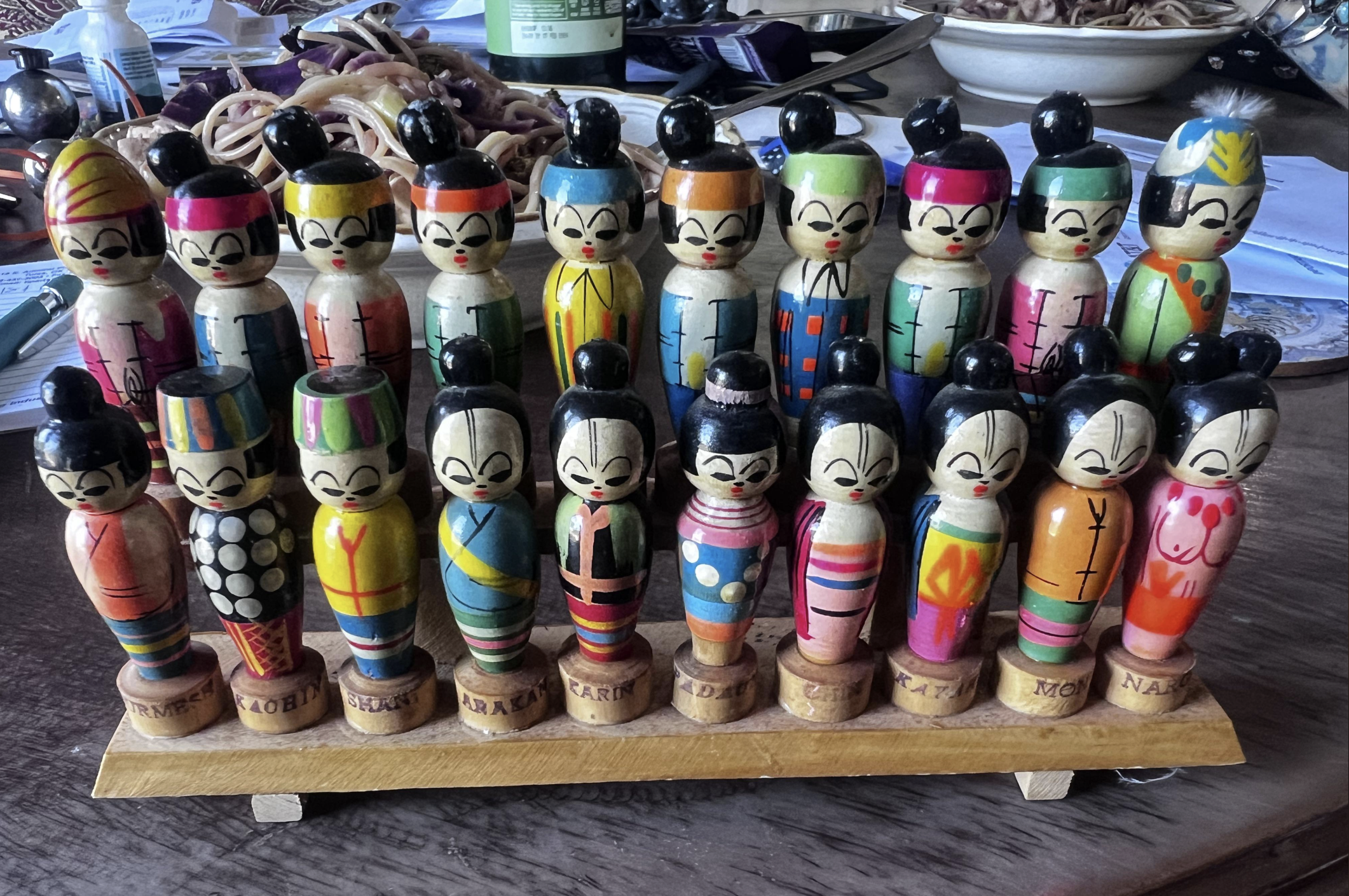 A set of colorful wooden dolls, each with unique painted outfits and hairstyles, standing on a wooden base with individual name labels. The background shows a table with papers, a bowl, and household items.