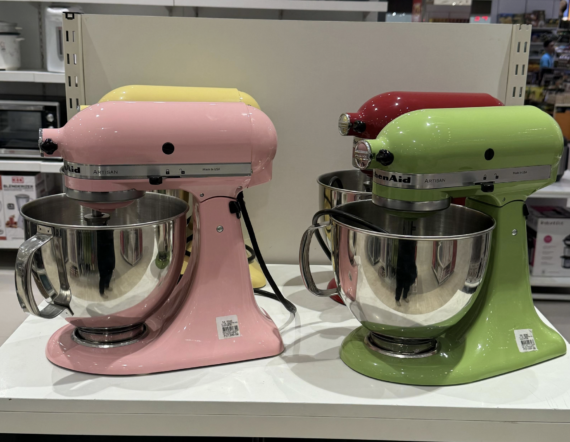 Four KitchenAid stand mixers in pastel colors&mdash;pink, yellow, green, and red&mdash;are displayed on a white shelf in a store, each with a stainless steel mixing bowl attached.