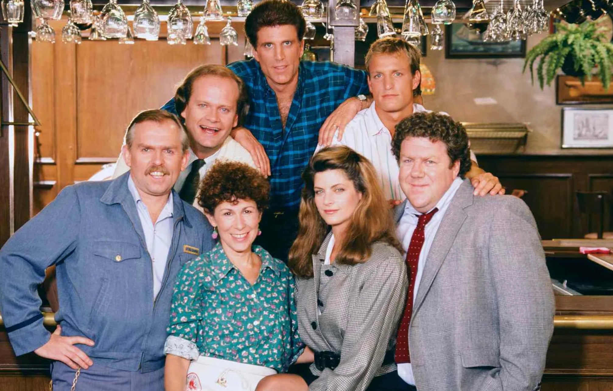 Seven people pose together behind and in front of a bar counter, with glasses hanging overhead and wooden shelves in the background. The group is smiling and dressed in casual and business attire.