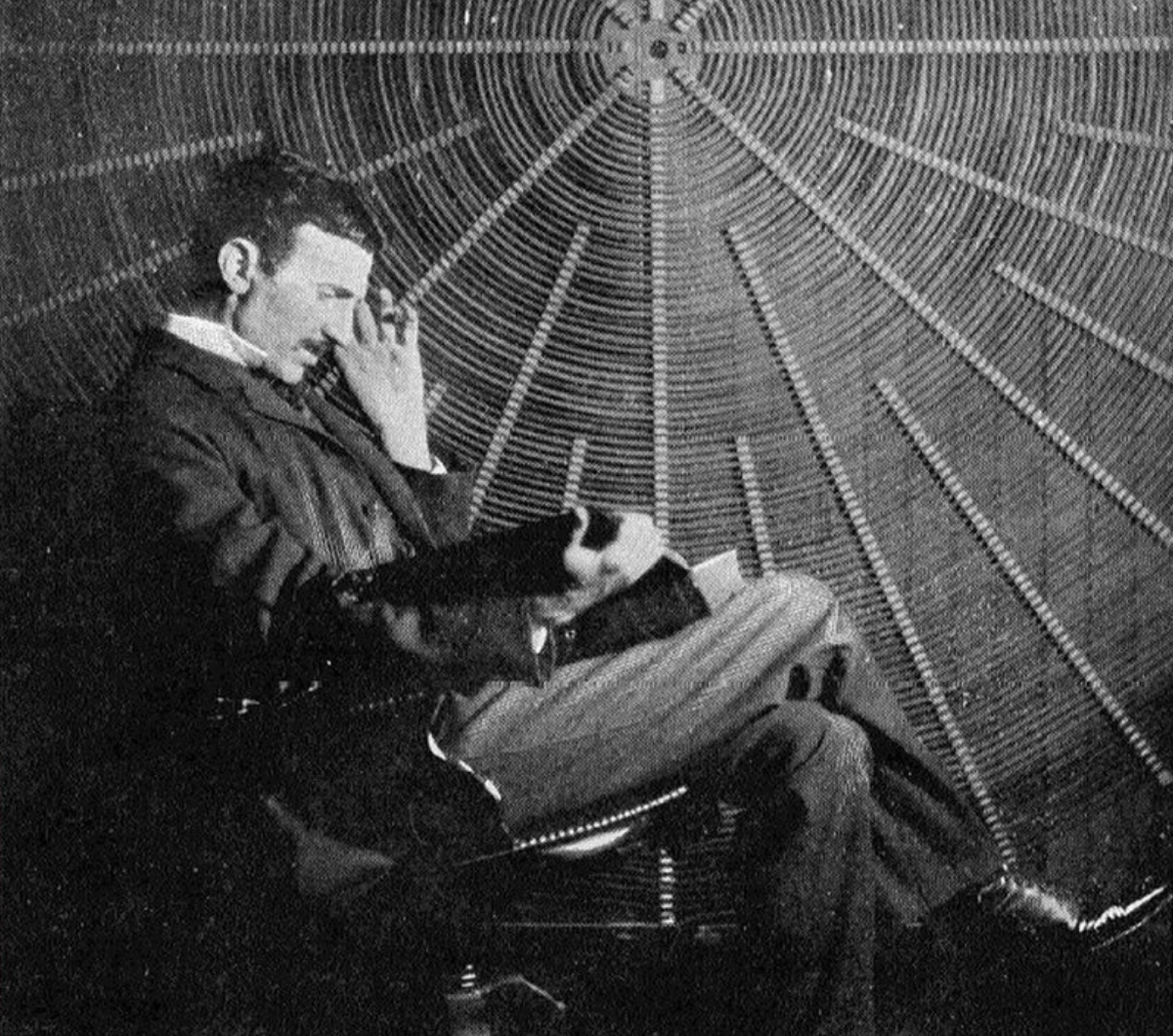 A man in a suit sits in a chair, reading papers, with a large, circular arrangement of wires or coils forming a dramatic pattern behind him. The scene appears monochrome and historical.
