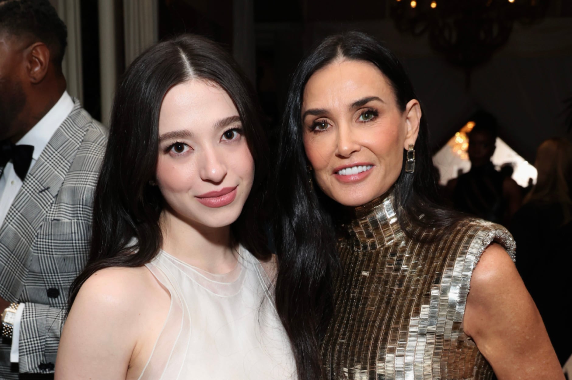 Two women pose together at an event. The woman on the left has long dark hair and wears a sleeveless white dress. The woman on the right has long dark hair and wears a sleeveless metallic gold dress. Other guests are visible in the background.