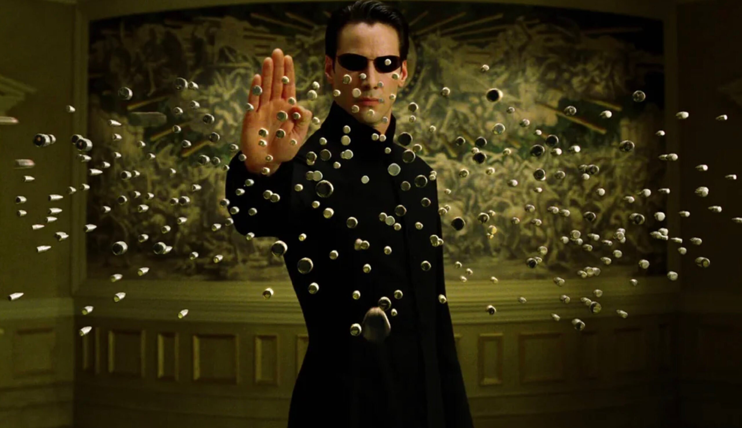 A man in a long black coat and sunglasses stands with one hand raised, stopping dozens of bullets in midair. The background is ornate and greenish, with blurred decorative details.