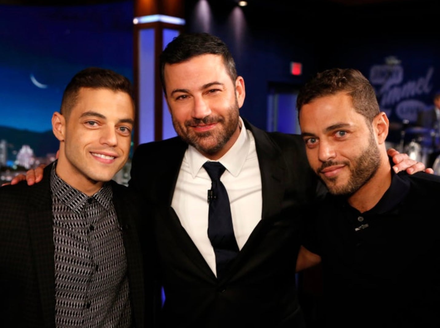 Three men stand closely together, smiling at the camera in a well-lit studio setting. The man in the middle wears a suit and tie, while the other two men wear casual shirts. The background has blue and purple tones.