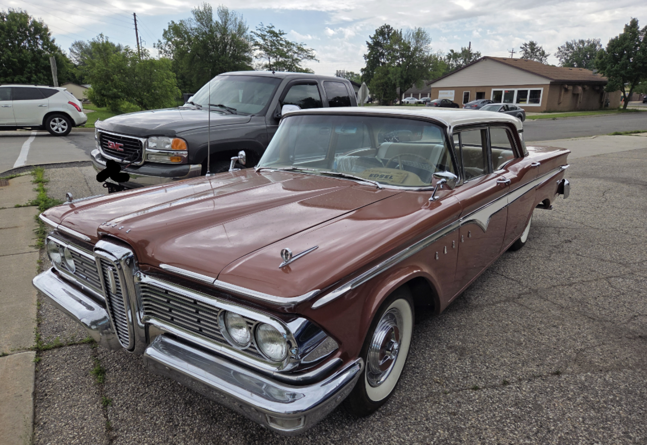 A vintage maroon and white classic car with chrome details is parked on a street near other vehicles and a single-story building in the background.
