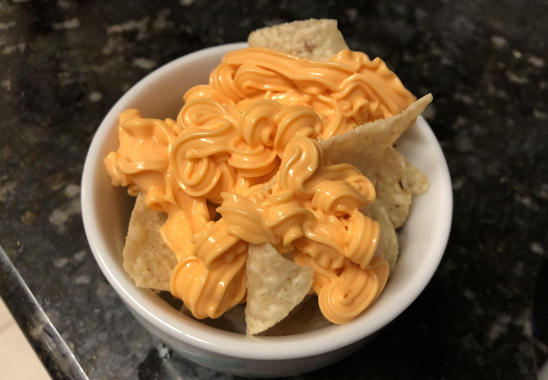 A white bowl filled with tortilla chips topped with a generous amount of orange cheese sauce, set on a dark countertop.