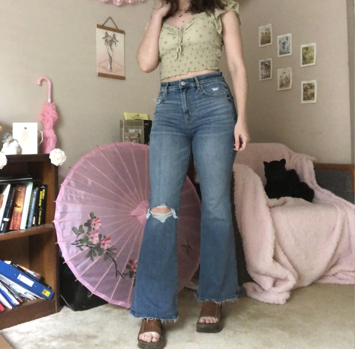 A woman in a green crop top, ripped blue jeans, and platform sandals stands indoors. Behind her are pink decorative umbrellas, bookshelves, and a black cat sitting on a pink blanket-covered chair.