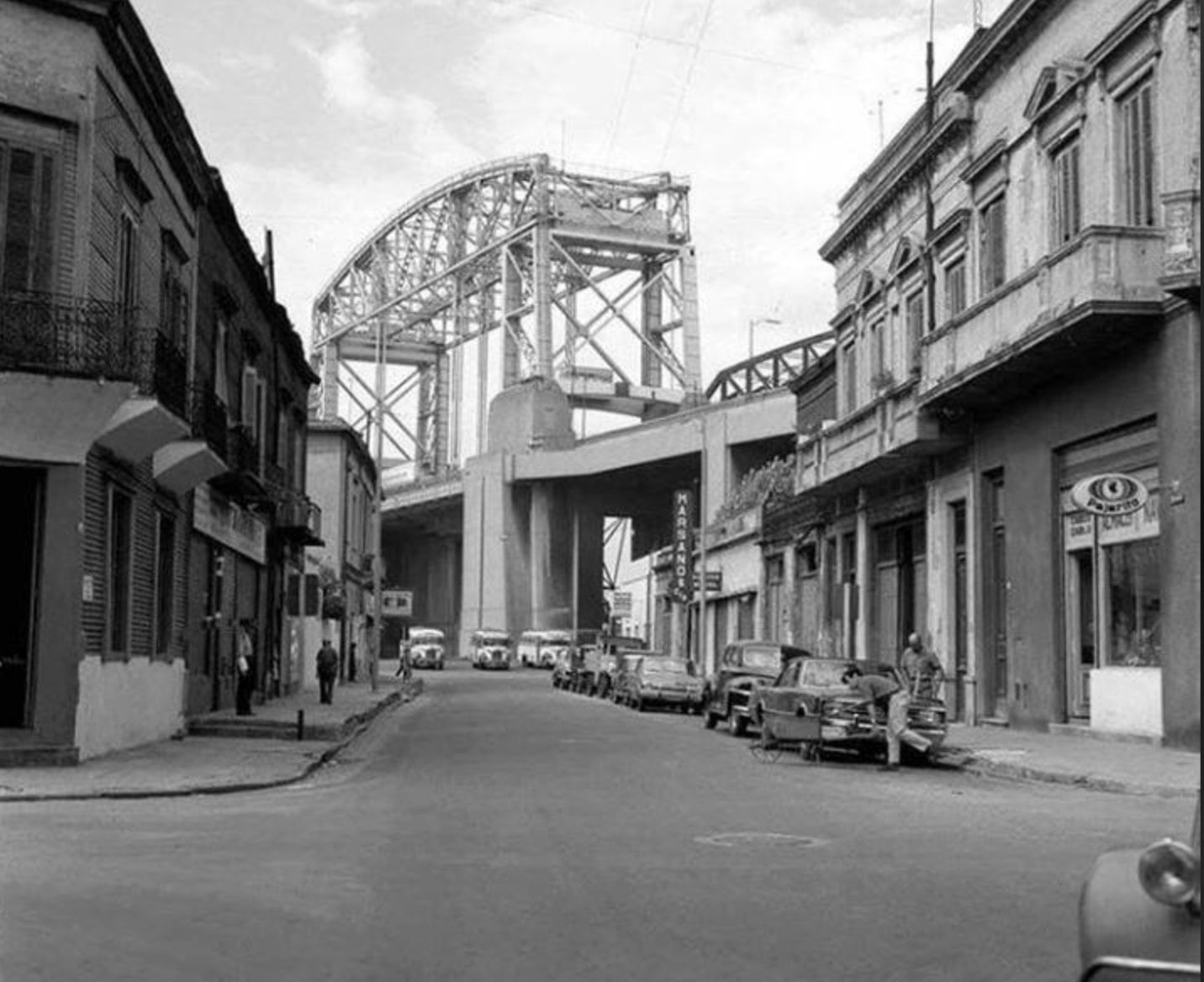 Black-and-white photo of a city street lined with old buildings and vintage cars, leading towards a large steel arch bridge in the background under a partly cloudy sky.