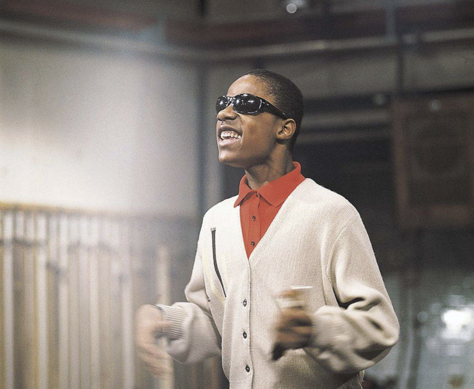 A young person wearing dark sunglasses, a beige cardigan, and a red collared shirt smiles while gesturing with their hands, standing indoors with blurred background elements.