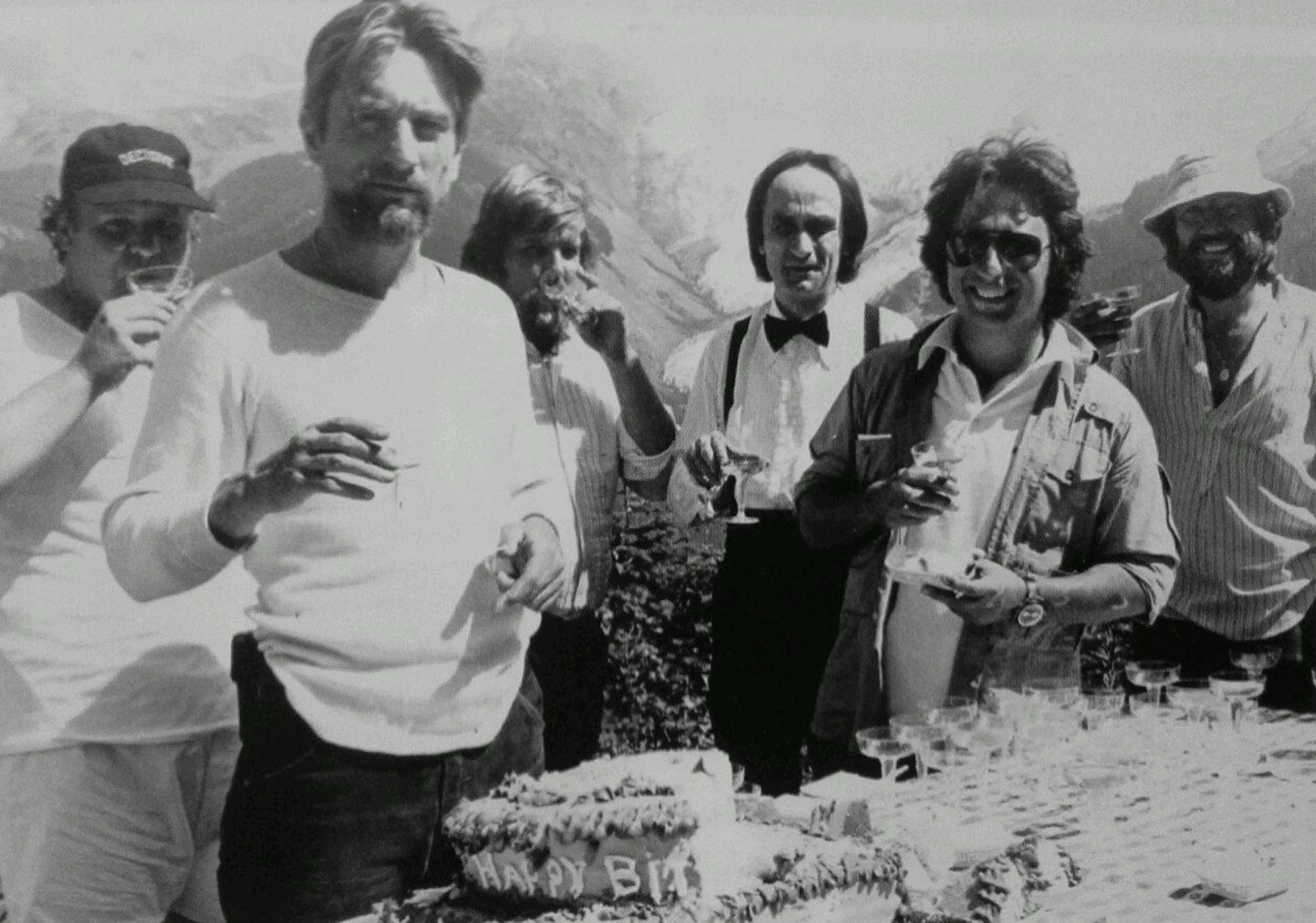 Six men stand outdoors by a table with a birthday cake and drinks, mountains in the background. Some smile or eat, one wears a bow tie, others wear casual clothes and sunglasses. The setting appears to be a celebration.