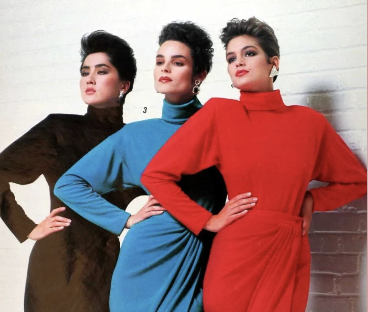 Three women stand side by side against a white wall, wearing high-neck, long-sleeve dresses in black, blue, and red. They pose confidently with hands on hips, embodying a bold 1980s fashion style.