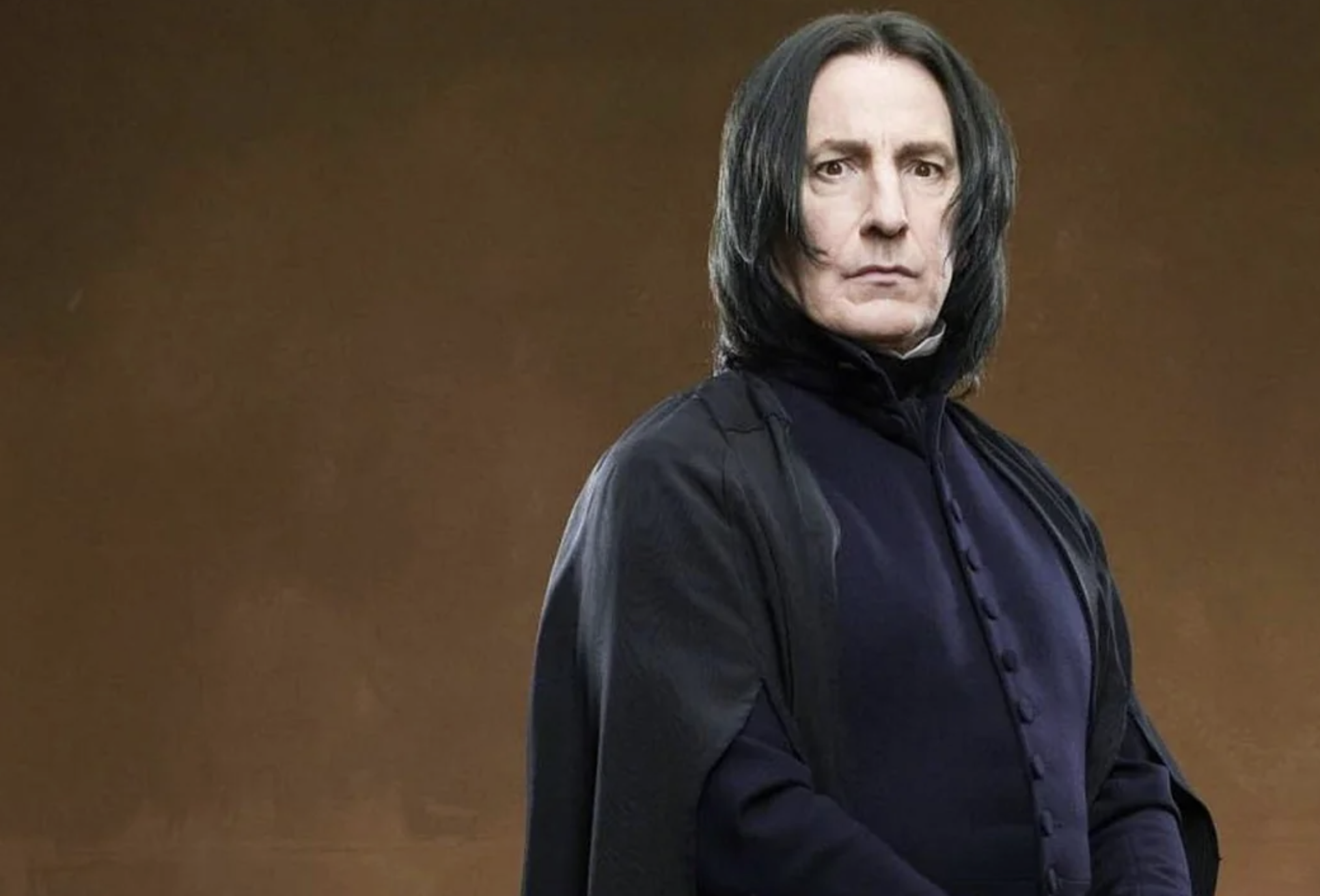 A person dressed in a long, black cloak and high-collared, dark clothing stands against a plain brown background, with shoulder-length black hair and a serious expression.