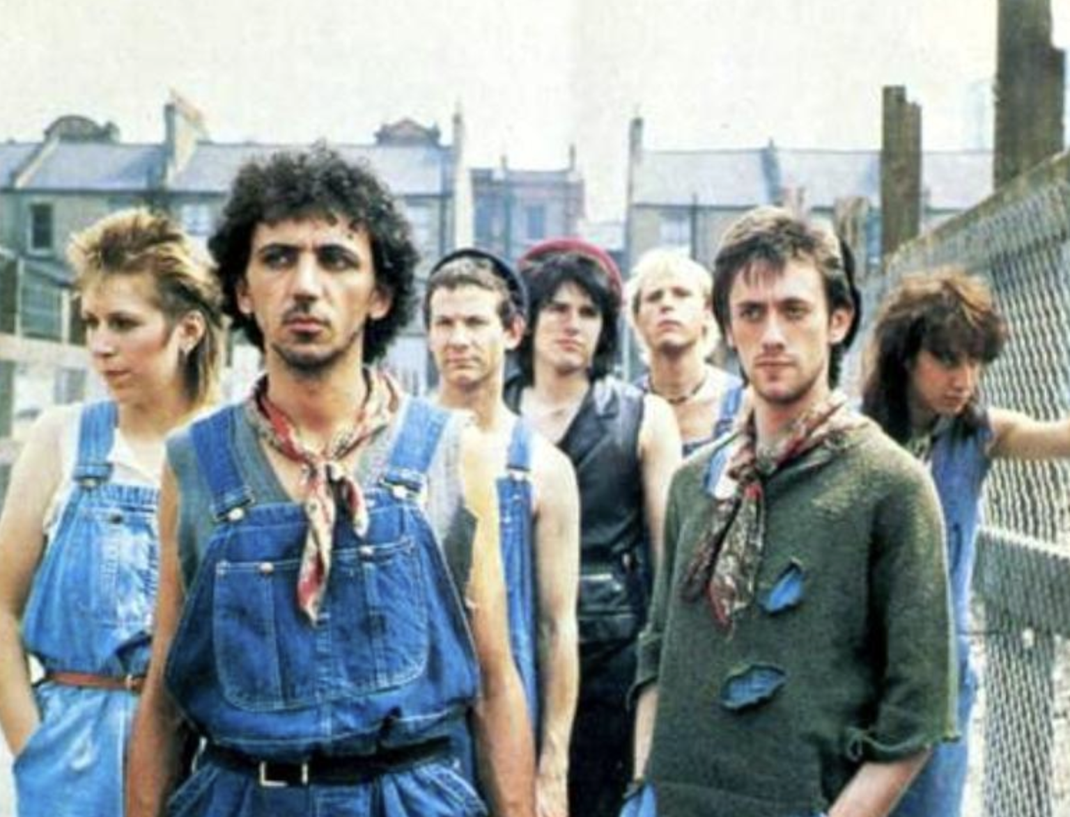 A group of seven young adults stand outdoors near a chain-link fence, wearing casual 1980s clothing like denim overalls and scarves. Buildings can be seen in the background.